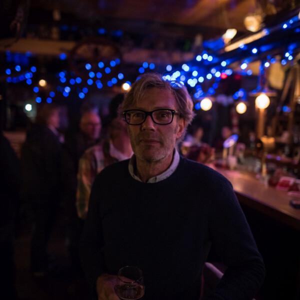Ein Mann mit Brille sitzt an der Bar in einer Kneipe und hält ein Glas in der Hand. 