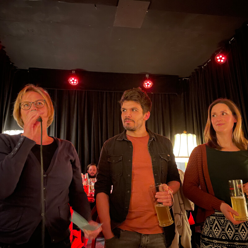 Drei Menschen stehen auf der Bühne: Eine Frau spricht ins Mikrofon, ein Man und eine Frau stehen daneben und schauen sie mit Bier in der Hand an.