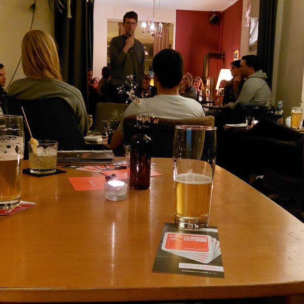 Ein Mann spricht ins Mikrofon vor Publikum. Im Vordergrund steht ein Bier neben dem Science goes Public Flyer.